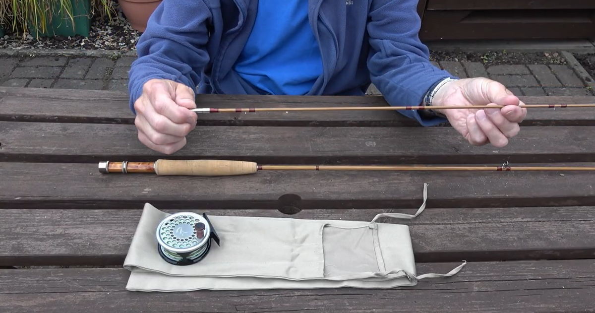 Angler holding a bamboo rod section above a reel and rod bag on a wooden table.